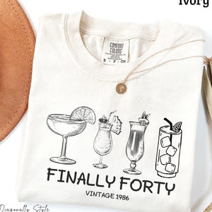 May include: An ivory t-shirt featuring a black line art graphic of four different cocktails and the text "FINALLY FORTY VINTAGE 1986". The shirt has a crew neckline and a Comfort Colors brand label on the inside collar, showing a size small.
