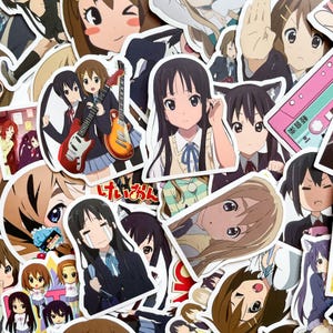 Pode incluir: Uma cole&ccedil;&atilde;o de autocolantes em estilo anime com v&aacute;rias personagens femininas. Os autocolantes mostram diferentes poses e express&otilde;es, alguns segurando guitarras ou usando uniformes escolares. A paleta de cores inclui tons de castanho, azul e rosa.