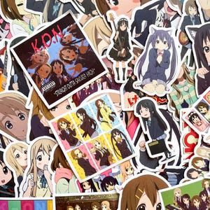 Pode incluir: Uma cole&ccedil;&atilde;o de autocolantes em estilo anime com v&aacute;rios personagens e designs. Alguns autocolantes incluem texto como "K.O.N" e "Straight Outta Sakura High". Os autocolantes exibem uma variedade de cores e poses, com foco na arte em estilo mang&aacute;.