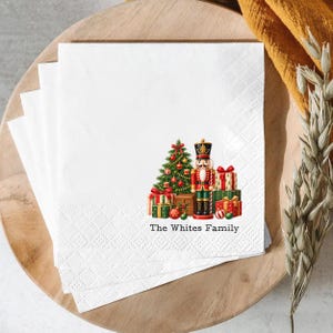 May include: A stack of white paper napkins featuring a printed illustration of a Christmas tree, a nutcracker, and wrapped gifts. The text "The Whites Family" is printed in black below the festive holiday scene on the square napkins.