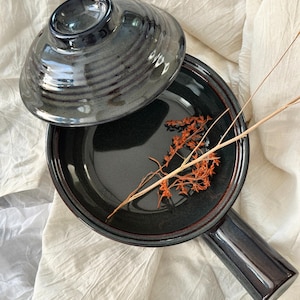 May include: A dark gray ceramic pan with a lid, featuring a handle and a slightly textured surface. The pan contains dried orange flowers. The lid is partially open, revealing the interior. The pan is on a white cloth.