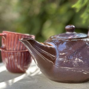 Op de afbeelding: Een bruine keramische theepot met bijpassende deksel en tuit. Twee gestapelde rode keramische kopjes op de achtergrond. De theepot en kopjes staan op een licht oppervlak, met een groene achtergrond.