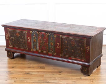 May include: A rectangular antique chest with a red base and a faded top. The chest features floral and geometric designs in gold, green, and brown. The chest is supported by short, curved legs.
