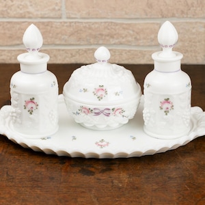 May include: A white vanity set with a tray, two bottles, and a lidded jar. The set is decorated with pink roses and purple accents. The bottles and jar have decorative stoppers.