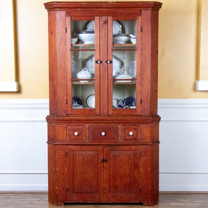 May include: A vintage corner cabinet with glass doors and a wooden base. The cabinet has a red-brown finish and features three drawers and two doors. The glass doors are open and reveal shelves filled with white dishes.