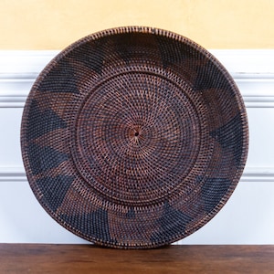May include: A large, round, woven basket with a dark brown and black pattern. The basket is made of natural materials and has a circular design.