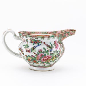May include: A white porcelain creamer with a floral and bird design in shades of pink, green, and blue. The creamer has a handle and a spout.
