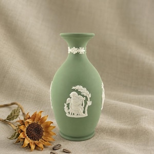 Op de afbeelding: Een matte saliegroene keramische vaas met witte reliëfversieringen. De hals is versierd met een bloemenband, terwijl het lichaam een centraal wit reliëf toont van een cherubijn die harp speelt onder een tak.