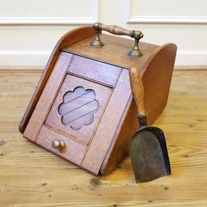 Op de afbeelding: Een houten kolenbak met een messing handvat en een bijpassende metalen schep. De kolenbak heeft een decoratieve uitsparing aan de voorkant en een klein lade.