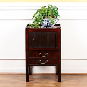 May include: A dark wood cabinet with two drawers and a single door, topped with a blue and white patterned planter filled with greenery. The cabinet has brass drawer pulls and a dark finish.