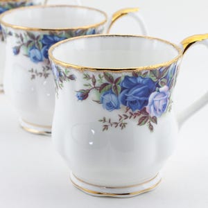 Op de afbeelding: Witte porseleinen mokken met een blauw rozenbloemmotief en gouden rand. De mokken hebben een gebogen handvat met een gouden accent. Het bloemmotief omvat blauwe en paarse rozen met groene bladeren.