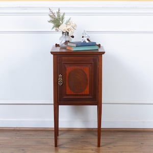 May include: A mahogany nightstand with a single door and a decorative oval inlay. The nightstand has a small brass handle and stands on four slender legs. A small vase of flowers and two books are on top.
