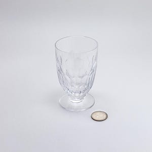 Op de afbeelding: Een helder glas met voet en een patroon. Het glas staat op een kleine, ronde voet. Een munt ligt naast het glas ter vergelijking. Het glas is waarschijnlijk om uit te drinken.