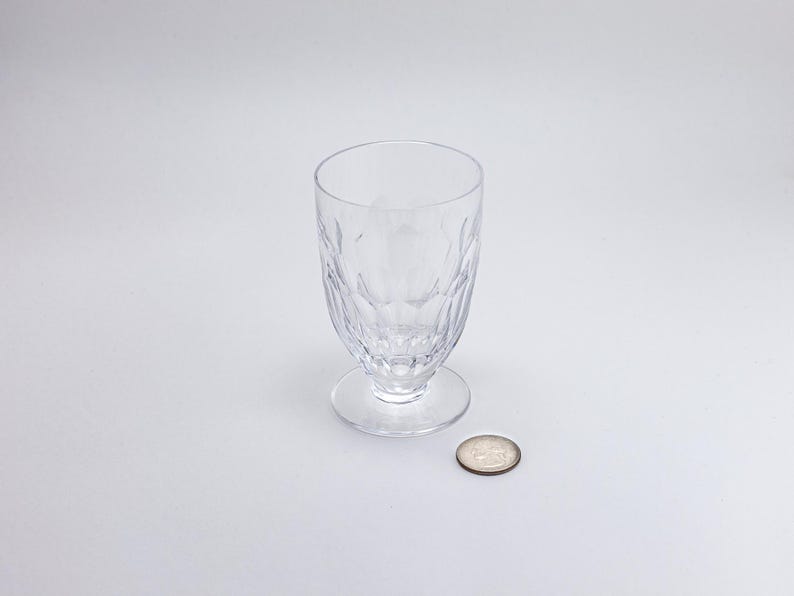 Op de afbeelding: Een helder glas met voet en een patroon. Het glas staat op een kleine, ronde voet. Een munt ligt naast het glas ter vergelijking. Het glas is waarschijnlijk om uit te drinken.
