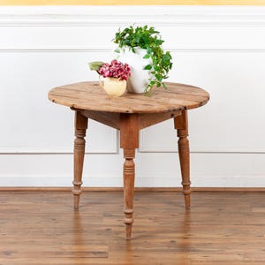 May include: A round, antique wooden table with a distressed finish. The table has a pedestal base with four turned legs. A white vase with green foliage and a small yellow pitcher with pink flowers sit on top.