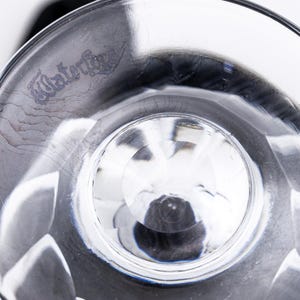 Op de afbeelding: Close-up van een helder, facetgeslepen glas met het woord "Waterford" ge&euml;tst op de rand. Het glas heeft een ronde basis en een gedetailleerd, gestructureerd ontwerp. Het glas wordt waarschijnlijk gebruikt om te drinken.