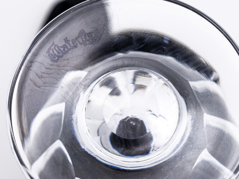 Op de afbeelding: Close-up van een helder, facetgeslepen glas met het woord "Waterford" ge&euml;tst op de rand. Het glas heeft een ronde basis en een gedetailleerd, gestructureerd ontwerp. Het glas wordt waarschijnlijk gebruikt om te drinken.