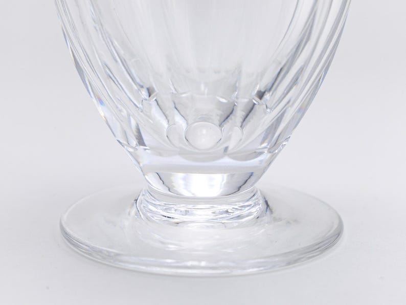 Op de afbeelding: Close-up van een helder glas met voet en een brede, cirkelvormige basis. Het glas heeft een uitlopende kom met verticale, getextureerde lijnen. De basis is een platte, cirkelvormige schijf. Het glas is transparant.