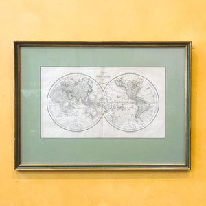 May include: Framed vintage world map print. The map is a detailed, two-hemisphere design in black and white, set against a light green mat. The frame is a dark gold color, and the artwork is displayed on a yellow wall.