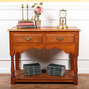 May include: A wooden console table with two drawers and a lower shelf. The table is made of oak and has a warm brown finish. It features decorative curved legs and a scalloped apron. The table is decorated with a bouquet of pink flowers, a brass clock, and a stack of books.