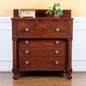 May include: A mahogany chest of drawers with four drawers and a top drawer with a hinged lid. The chest has turned legs and crystal knobs. A small decorative box sits on the top of the chest.