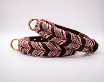 May include: Two braided dog collars. The collars feature a combination of brown, pink, and lavender fabric with gold-colored accents. Each collar has a gold-colored metal ring. The collars are set against a white background.