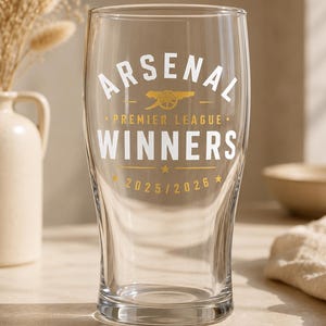May include: A clear pint glass with the text "ARSENAL PREMIER LEAGUE WINNERS 2025/2026" in white and gold lettering. The glass is curved and suitable for beer or other drinks. The glass is approximately 15 cm tall.