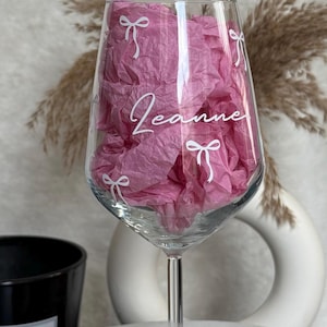 May include: A clear wine glass with a long stem, filled with pink tissue paper. The glass is decorated with white bow designs and the name "Jeanne" in cursive. A black candle and decorative vase are in the background.