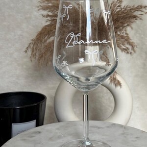 May include: A clear wine glass with a long stem and a round base. The glass is decorated with white bow designs and the name "Jeanne" in cursive. The glass is on a marble surface.