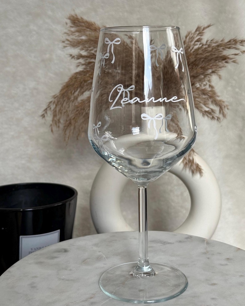 May include: A clear wine glass with a long stem and a round base. The glass is decorated with white bow designs and the name "Jeanne" in cursive. The glass is on a marble surface.