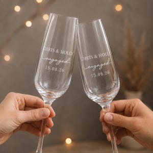 May include: Two clear glass champagne flutes featuring white etched text that reads CHRIS & HOLLY engaged 15.08.26. The tall, slender glasses have a smooth, transparent finish and are held up to create a celebratory toast.