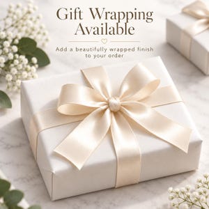 May include: A square gift box wrapped in plain white paper with a large, centered cream-colored satin ribbon bow. The text reads Gift Wrapping Available and Add a beautifully wrapped finish to your order with a small heart icon.
