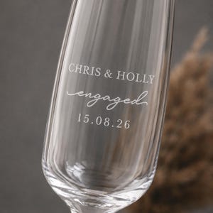 May include: A clear glass champagne flute featuring an etched design with the text CHRIS & HOLLY, the word engaged in a cursive script, and the date 15.08.26. The glass has a vertical ribbed texture along the lower portion of the bowl.