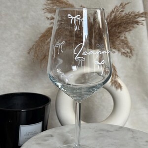 May include: A clear wine glass with a long stem, decorated with white bow illustrations and the name "Jeanne" in cursive. The glass is on a marble surface, with a black candle and decorative vase in the background.