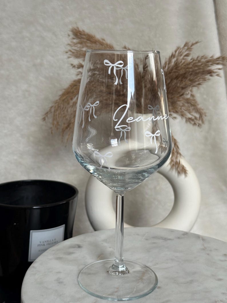 May include: A clear wine glass with a long stem, decorated with white bow illustrations and the name "Jeanne" in cursive. The glass is on a marble surface, with a black candle and decorative vase in the background.