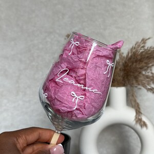 May include: A clear wine glass with the name "Leanne" in white script, adorned with white bow designs. The glass is filled with pink tissue paper. The glass is held by a person.