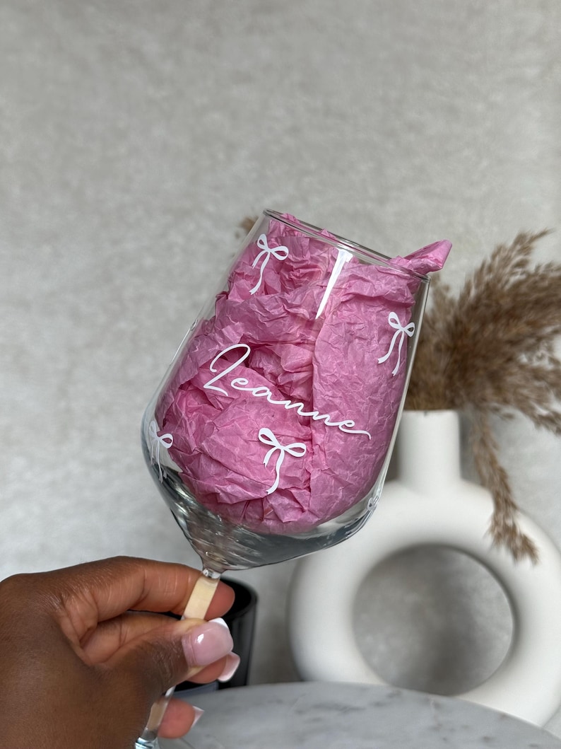 May include: A clear wine glass with the name "Leanne" in white script, adorned with white bow designs. The glass is filled with pink tissue paper. The glass is held by a person.