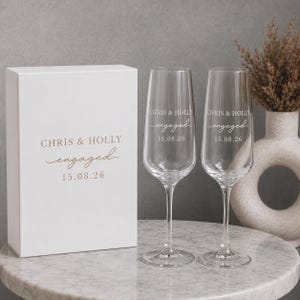 May include: A white rectangular gift box and two clear glass champagne flutes. The box and glasses are printed with the text CHRIS & HOLLY, engaged, and the date 15.08.26 in a gold-colored font. The flutes have tall stems and bases.
