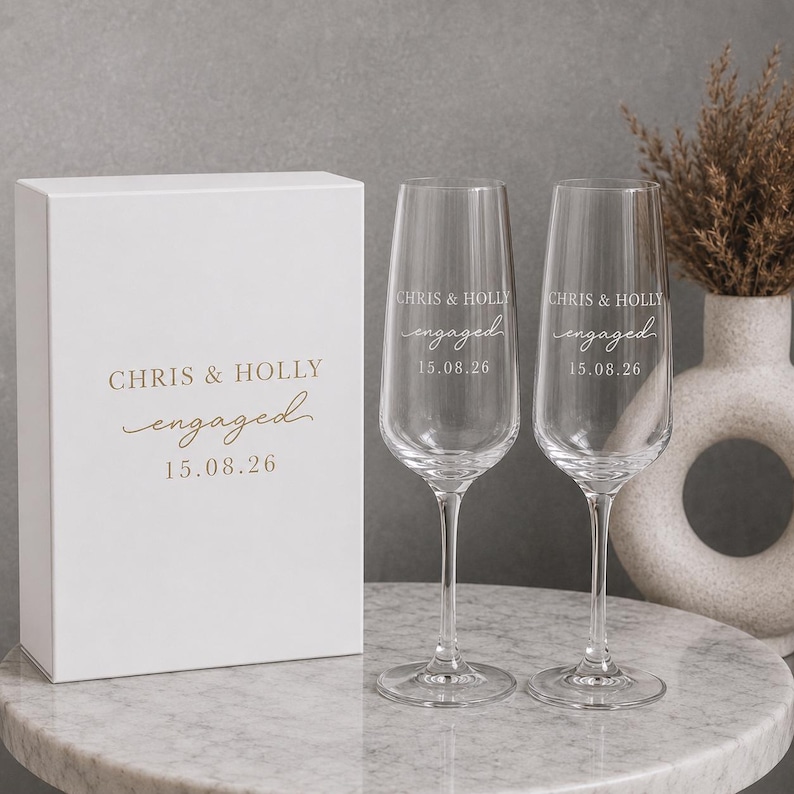May include: A white rectangular gift box and two clear glass champagne flutes. The box and glasses are printed with the text CHRIS & HOLLY, engaged, and the date 15.08.26 in a gold-colored font. The flutes have tall stems and bases.