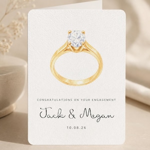 May include: A white engagement greeting card featuring a watercolor illustration of a gold ring with a round diamond. The text reads: CONGRATULATIONS ON YOUR ENGAGEMENT, Jack & Megan, and the date 10.08.26 in black ink on the front.