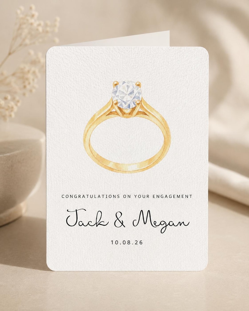 May include: A white engagement greeting card featuring a watercolor illustration of a gold ring with a round diamond. The text reads: CONGRATULATIONS ON YOUR ENGAGEMENT, Jack & Megan, and the date 10.08.26 in black ink on the front.