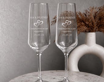May include: Two clear glass champagne flutes featuring an etched design with the names Sam & Heather, two interlocking hearts, the text Congratulations on your engagement, and the date 16.06.26 centered on the front of each glass.
