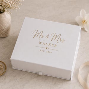 May include: A white gift box with a pull tab, featuring gold script that reads "Mr & Mrs WALKER" with a heart and "EST. 2026". The box is on a neutral surface, suggesting a wedding or anniversary theme.