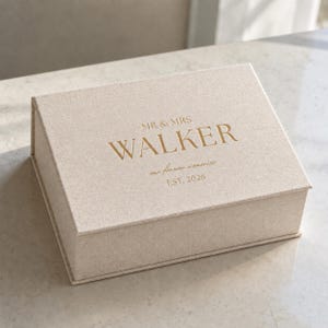 May include: A rectangular beige linen-textured keepsake box with a hinged lid. The top features gold foil lettering that reads MR & MRS WALKER, our forever memories, and EST. 2026, centred in a clean, serif and script font combination.