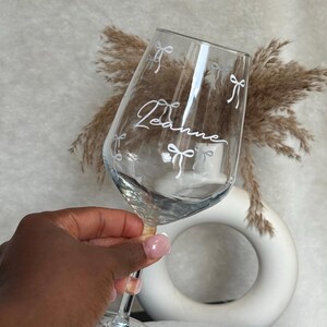 May include: A clear wine glass with the name "Leanne" in cursive script and white bow accents. The glass is held up, showcasing the design. The stem is clear with a small pink bead detail.
