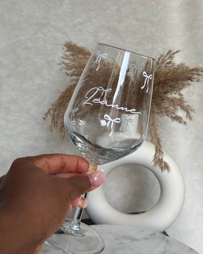 May include: A clear wine glass with the name "Leanne" in cursive script and white bow accents. The glass is held up, showcasing the design. The stem is clear with a small pink bead detail.