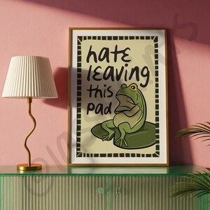 May include: A framed art print with a cartoon frog on a lily pad. The green frog has yellow eyes and is on a green lily pad. The print has the text "hate leaving this pad".