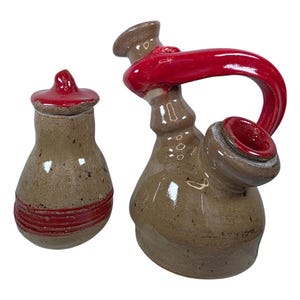 Puede incluir: Dos piezas de cerámica hechas a mano. Una es un pequeño tarro marrón con tapa roja y una franja roja. La otra es una pipa de agua marrón y curvada con asa y detalles rojos. Ambas piezas tienen un aspecto moteado y rústico.