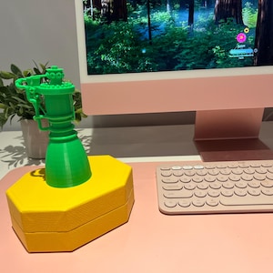 May include: A bright green, 3D-printed rocket-shaped object is displayed on a yellow, hexagonal base. The object is set against a backdrop of a computer monitor, keyboard, and a small potted plant.