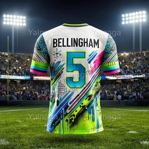 Może przedstawiać: Biała koszulka z napisem "BELLINGHAM" i numerem "5" w kolorze turkusowym. Koszulka ma kolorowy design z neonowymi zielonymi, niebieskimi i różowymi akcentami. Rękawy mają wielokolorowy wzór w paski. Koszulka znajduje się na zielonym boisku.